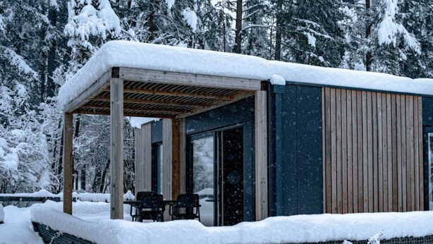 Eine moderne Holzhütte mit überdachter Terrasse, einem Essbereich und einem großen Fenster.