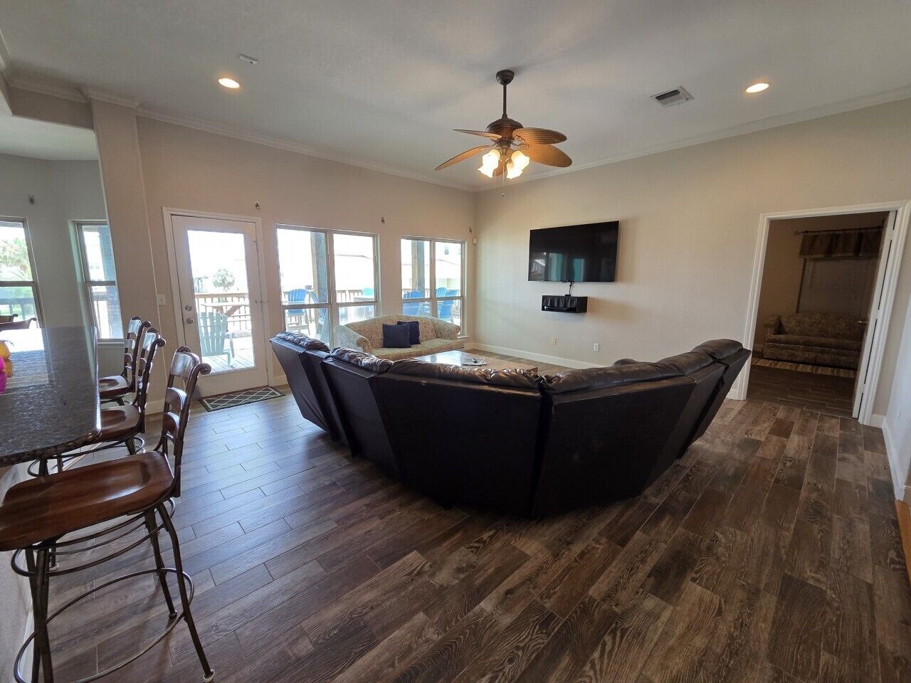 A spacious living area with a large dark-colored sectional sofa, a dining table with chairs, a ceiling fan, and a flat-screen TV mounted on the wall.