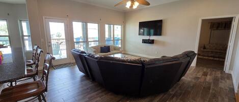 A spacious living area with a large dark-colored sectional sofa, a dining table with chairs, a ceiling fan, and a flat-screen TV mounted on the wall.