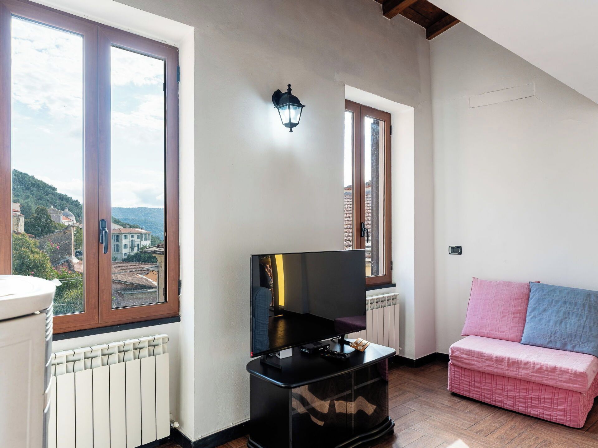 A room with a television, a radiator, a sofa, and a window with a view of buildings.