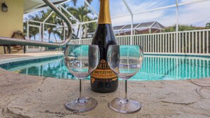 Una botella de champán Brut Cuvée y dos copas sobre una superficie de piedra junto a una piscina con una cerca blanca y palmeras al fondo.