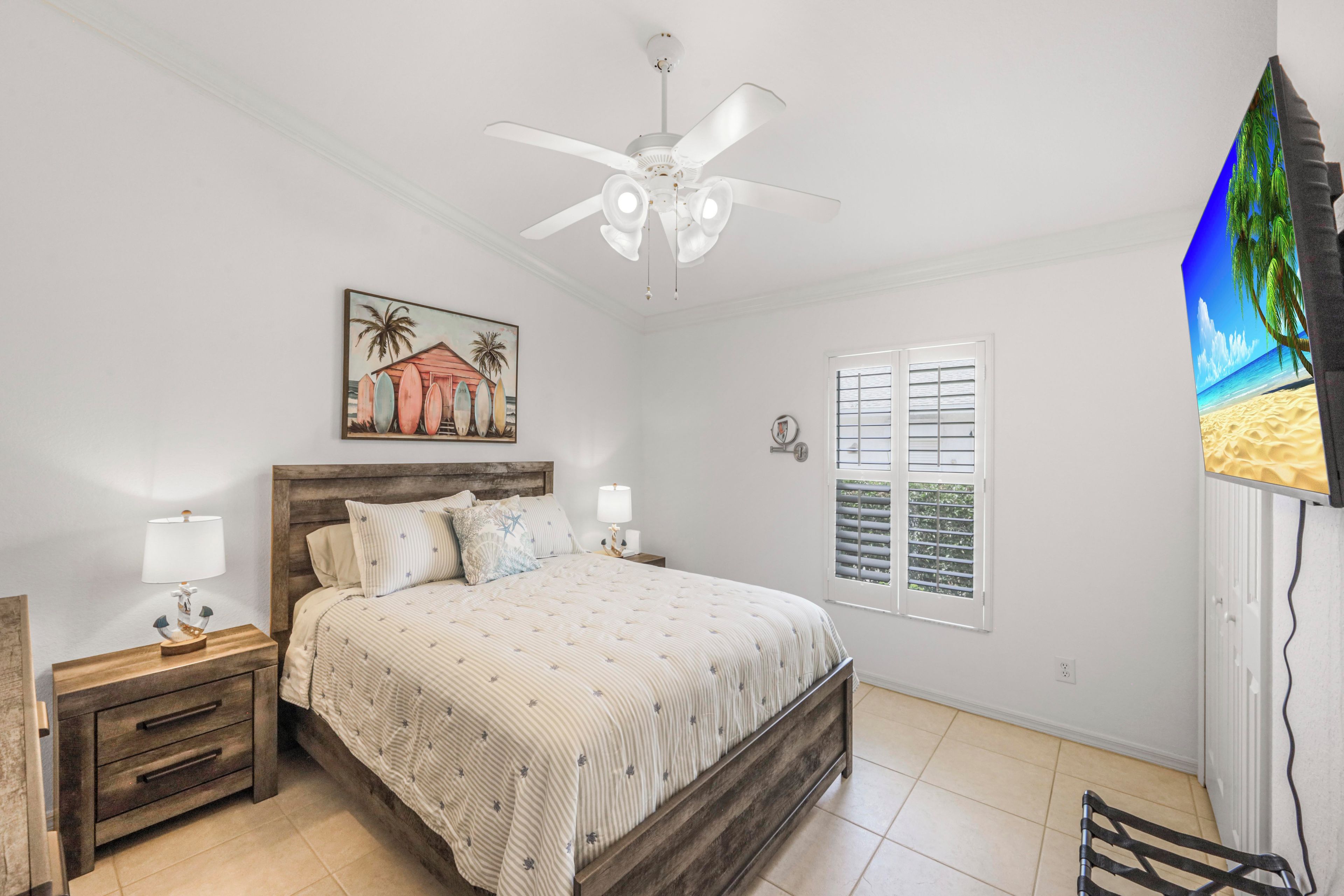 A bedroom with a bed, nightstands, a ceiling fan, a window with shutters, and a painting on the wall.
