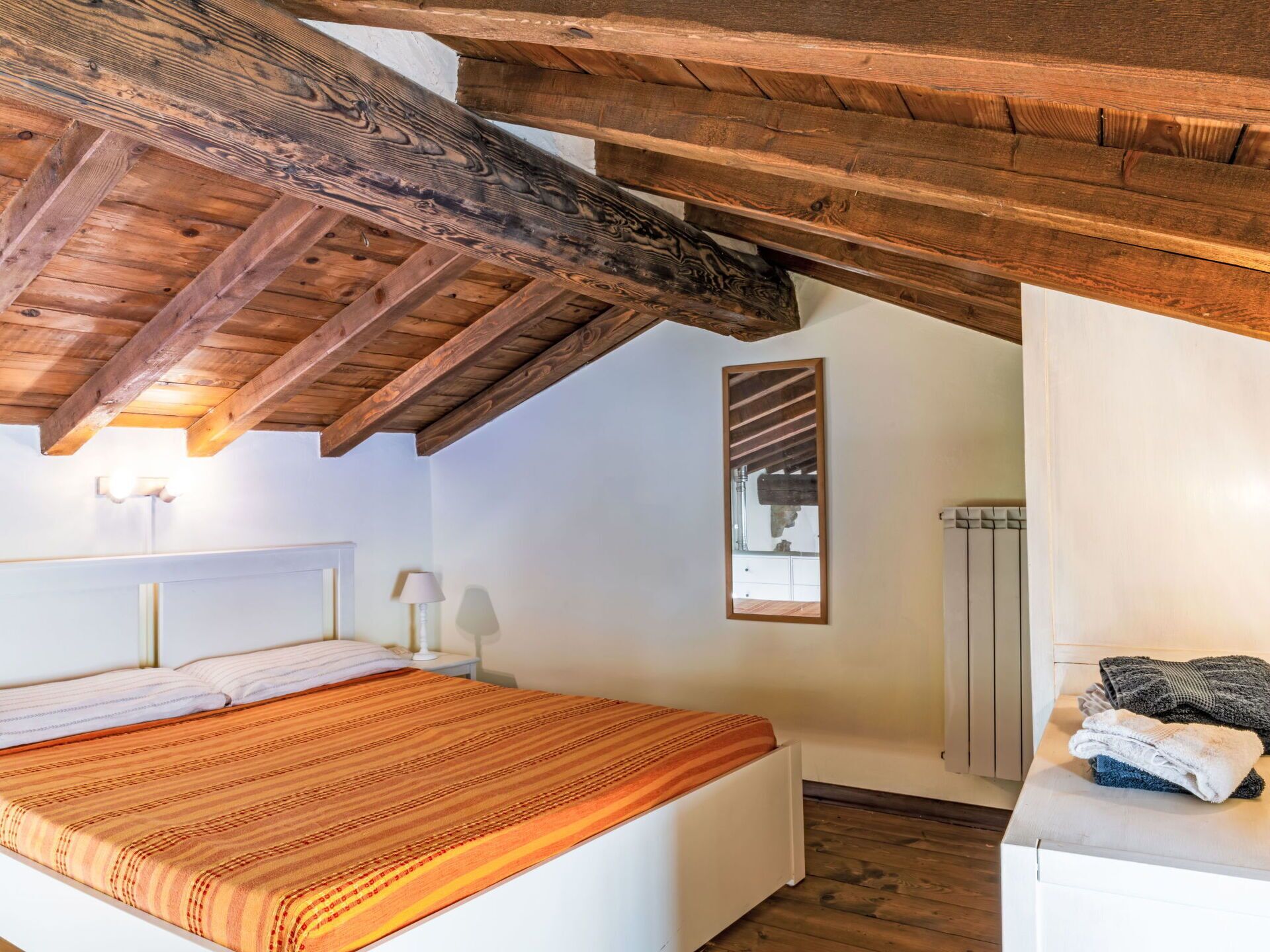 A bedroom with a wooden ceiling, a bed with a striped blanket, a bedside table with a lamp, and a radiator.