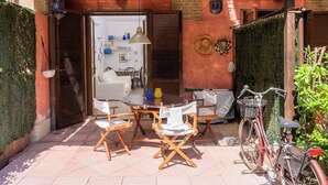 Zona exterior amb mobiliari, taula, cadires, una bicicleta i l'entrada d'un edifici.