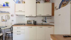 Una cuina amb armaris blancs, fornet a gas, un rentapeus i una petita zona de dinar amb una cadira i taula.