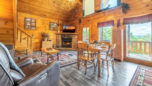 L’intérieur d’une cabane en rondins, avec une cheminée, des meubles en bois et une grande fenêtre donnant sur un paysage verdoyant.