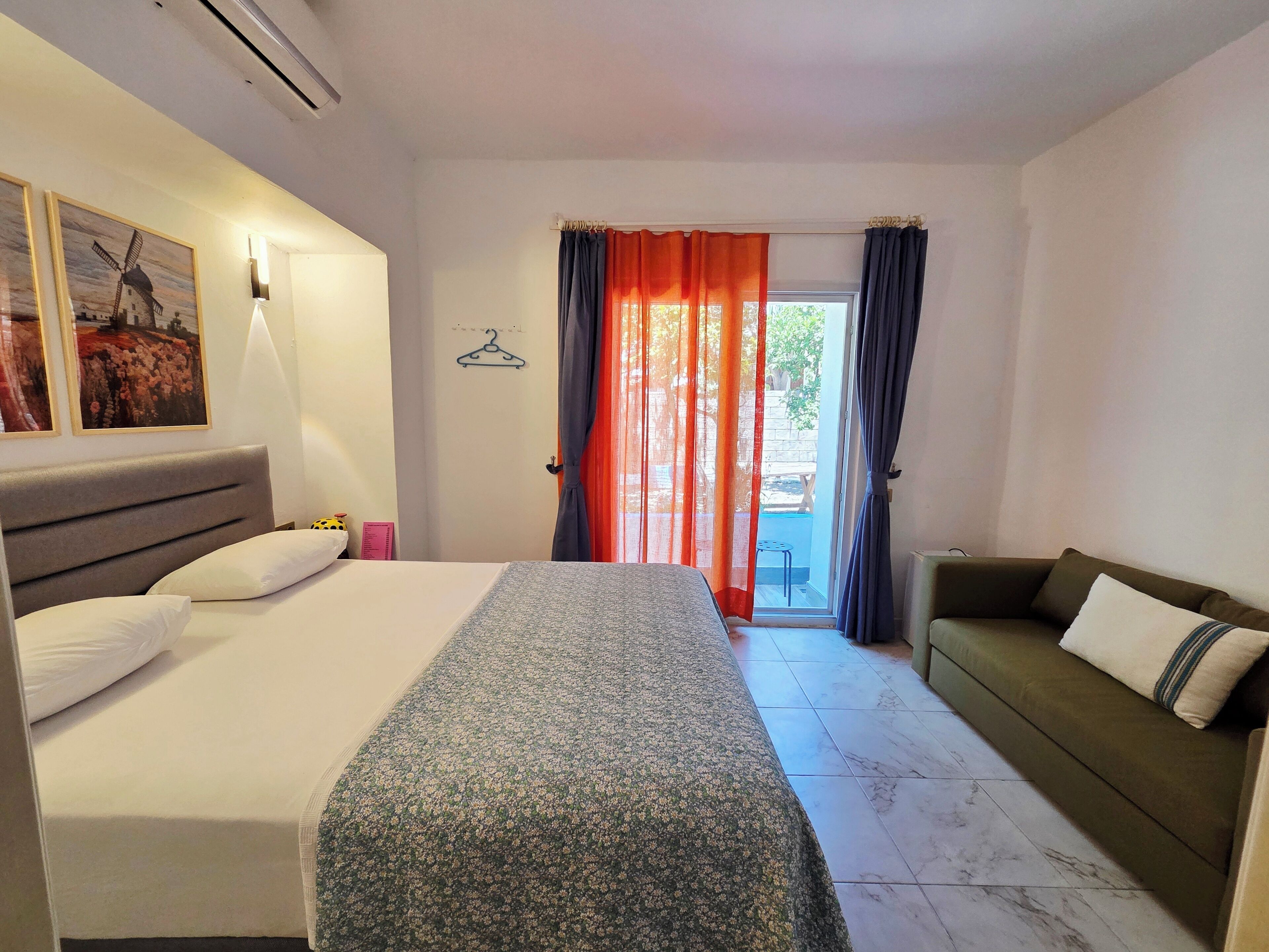 Quarto de hotel com cama, sofá, janela com cortinas, luminária na parede e porta com cabide.