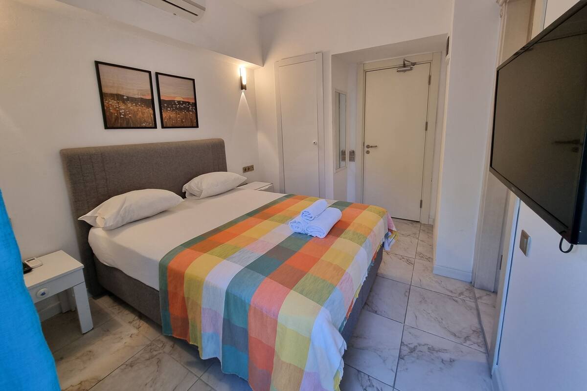 A hotel room with a bed, a bedside table, a TV, and a door leading to another room.