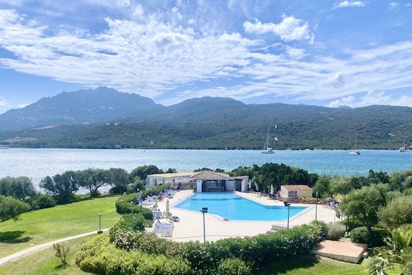 A swimming pool surrounded by greenery, a building with a tiled roof, and a view of a lake with boats and mountains in the background. - P.Rotondo1 B106 S, two-room apartment with 5 beds (Porto Rotondo)