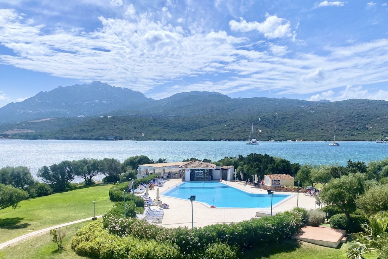 Ein Schwimmbecken, umgeben von viel Grün, ein Gebäude mit Ziegeldach und im Hintergrund ein See mit Booten und Bergen.