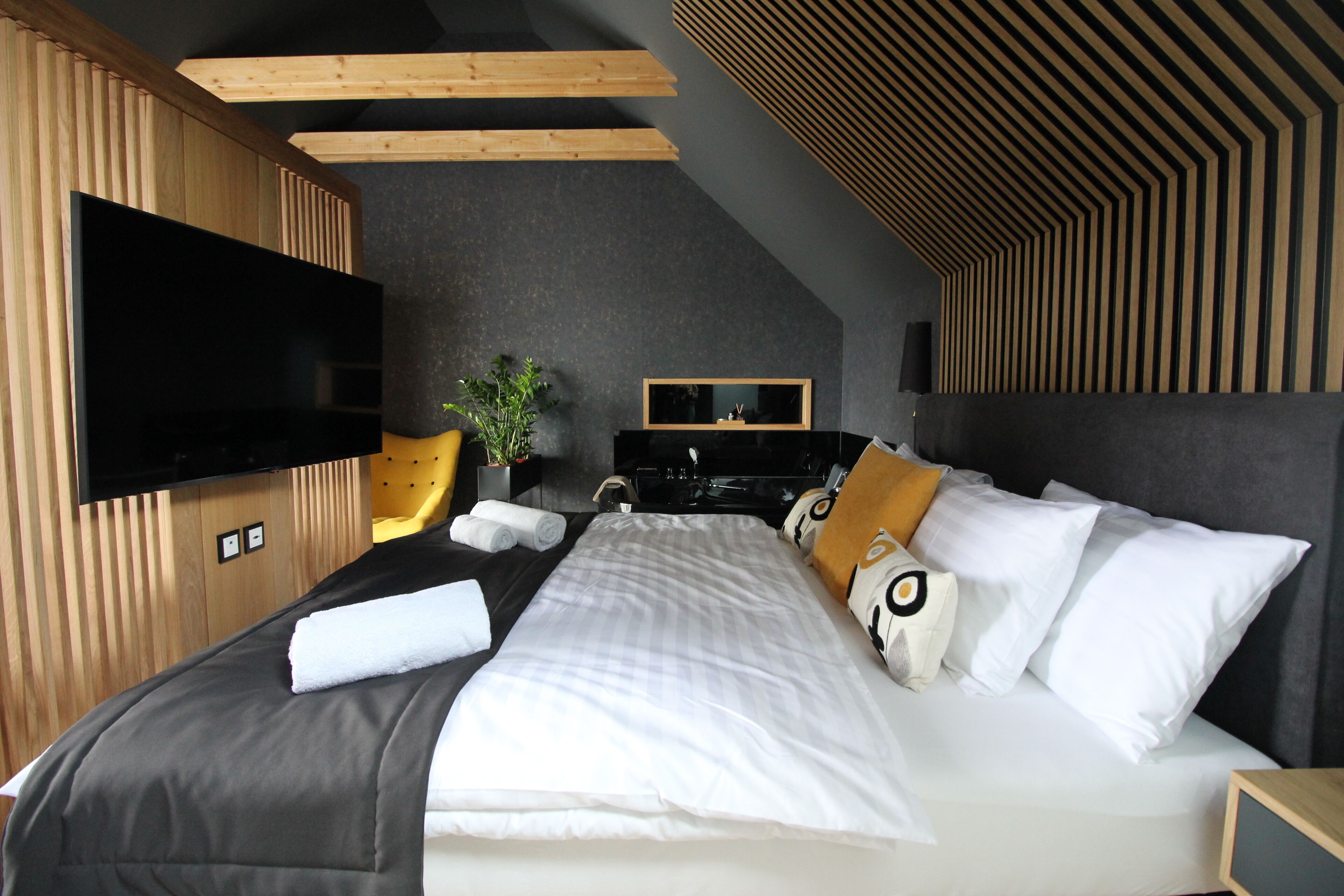 A modern hotel room with a flat-screen TV, a bed with white and patterned pillows, a wooden headboard, and a dark ceiling.