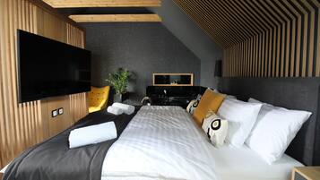 A modern hotel room with a flat-screen TV, a bed with white and patterned pillows, a wooden headboard, and a dark ceiling.