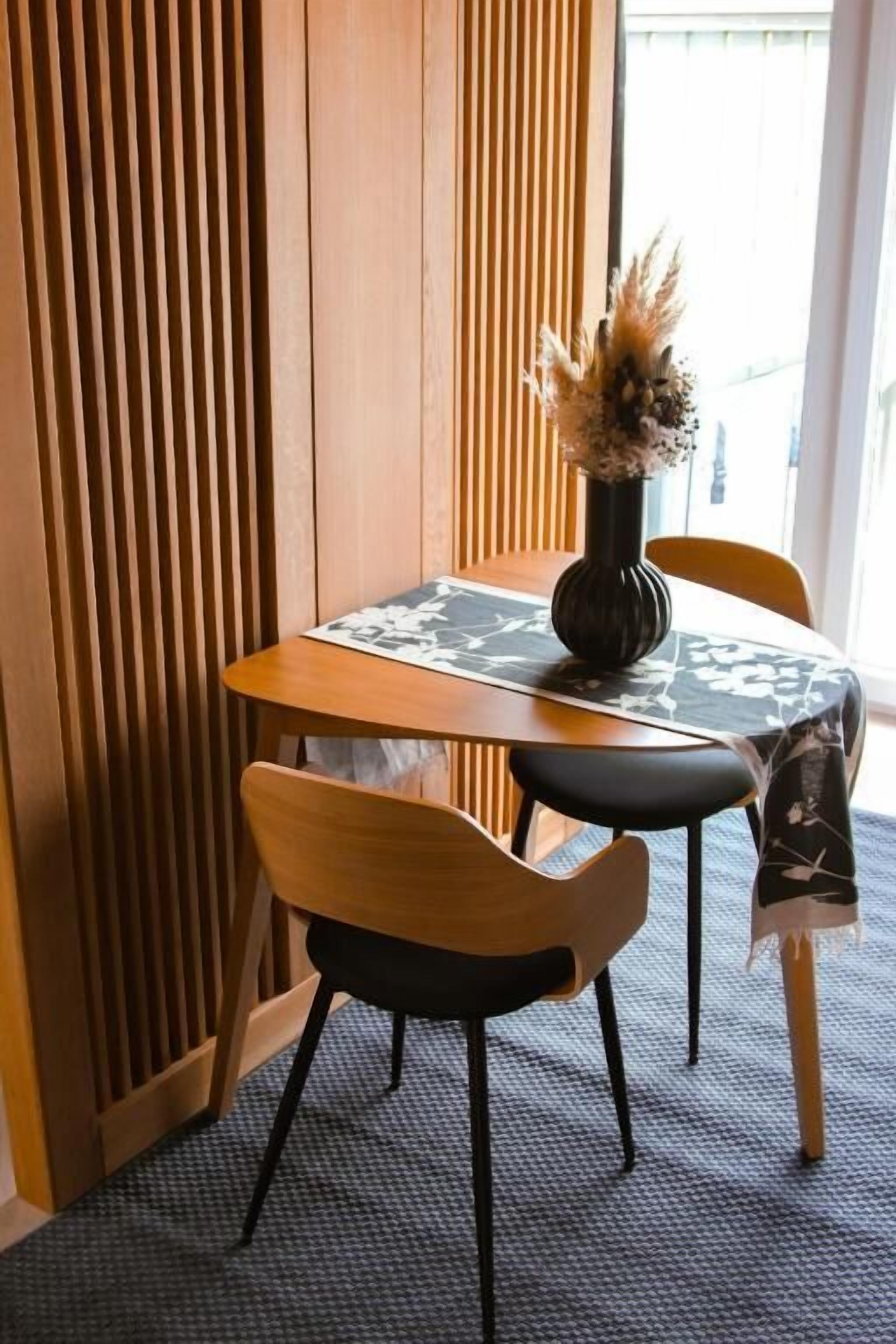 Ein Essbereich mit Tisch, Stühlen, einer Vase mit Trockenblumen und einer gemusterten Tischdecke.