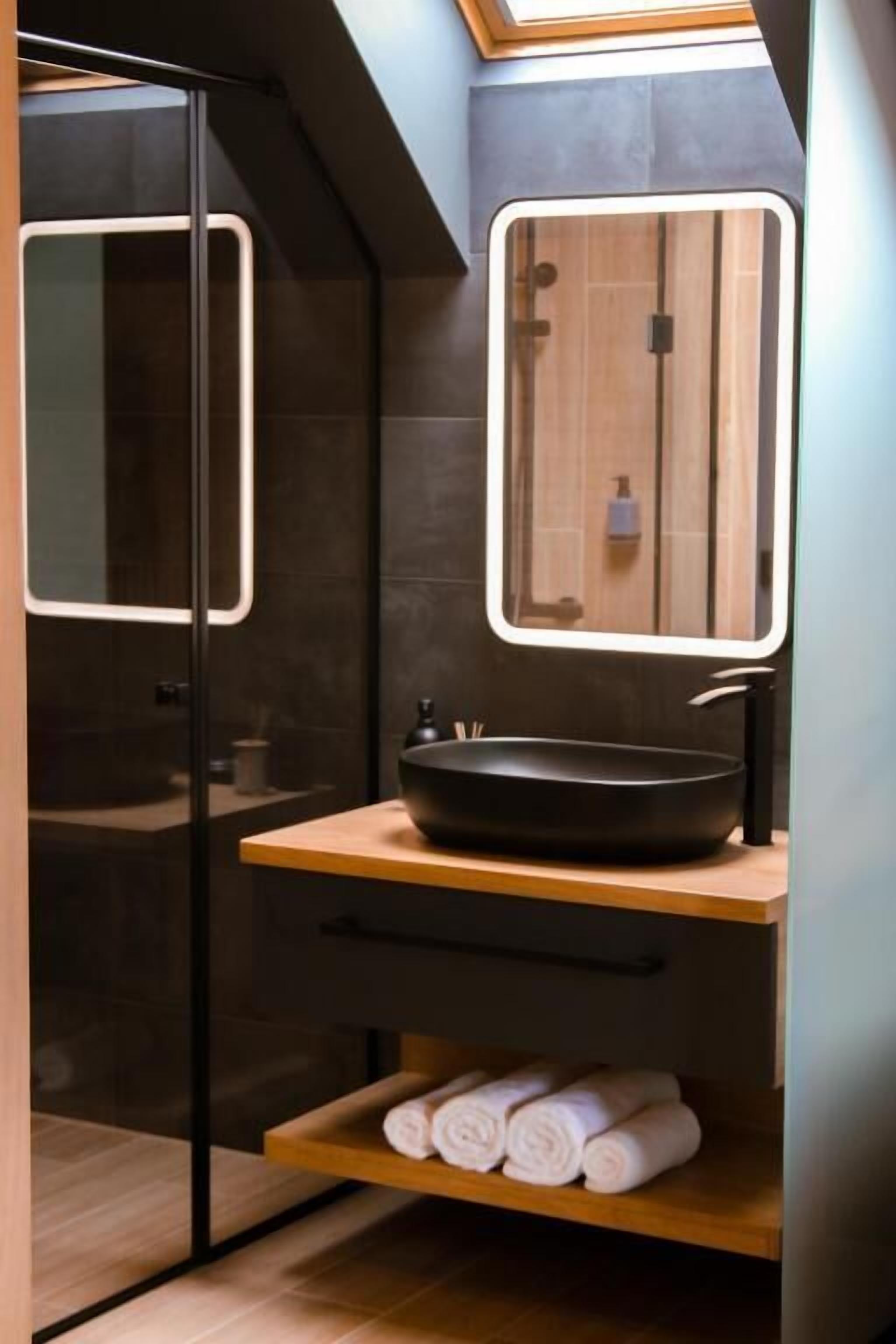 Ein modernes Badezimmer mit einer Duschkabine aus Glas, einem Holzwaschtisch mit schwarzem Becken und einem Oberlicht über dem Spiegel.