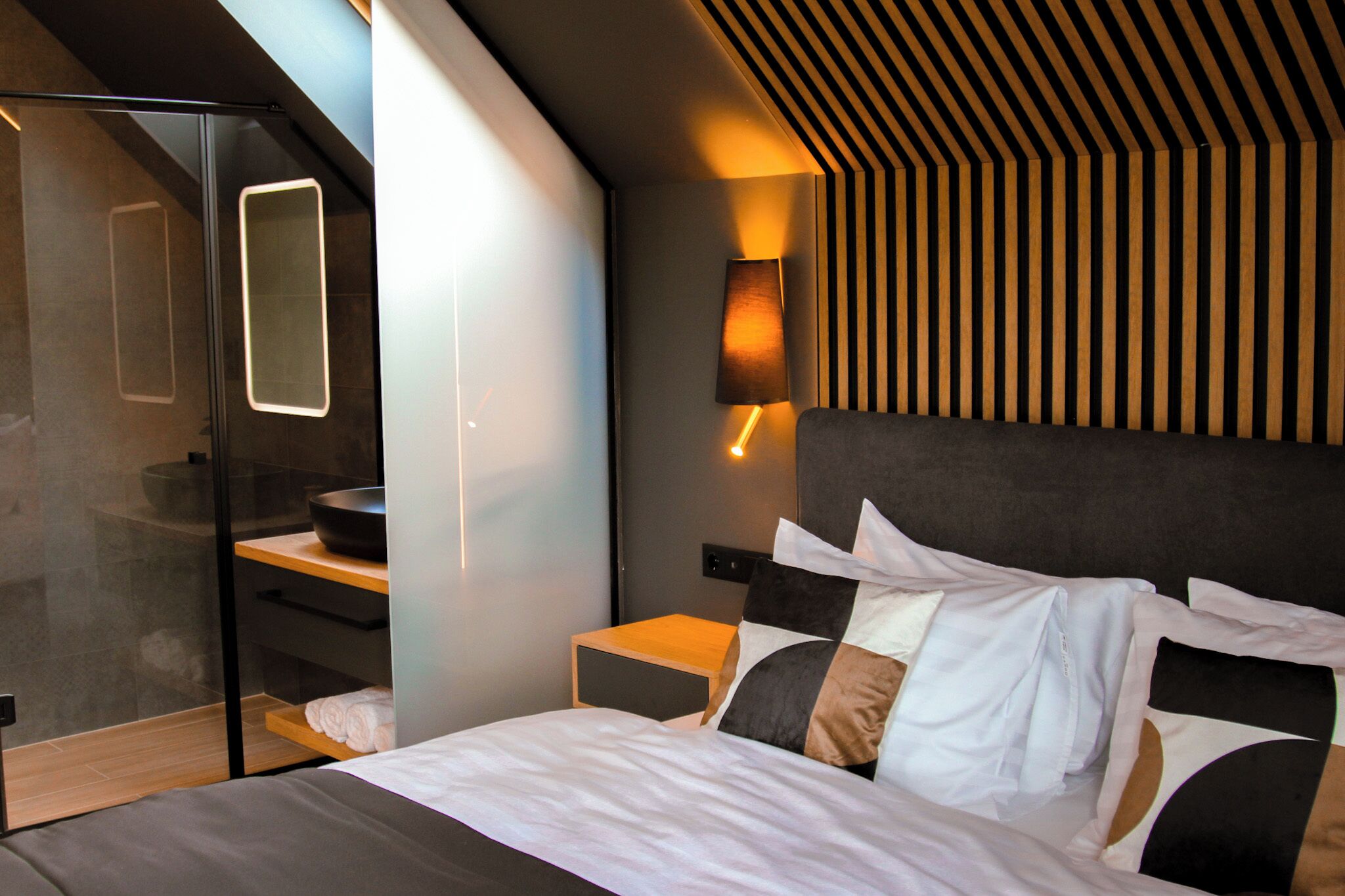 A modern bedroom with a bed, bedside tables, a built-in bench, a glass shower enclosure, and a sloped ceiling with wooden paneling.