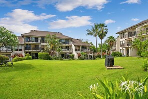 A courtyard with a lawn, a barbecue grill, and a building with balconies and windows. - Aston Poipu Kai | Walk to Beach | Pools | Garden Views (Koloa)