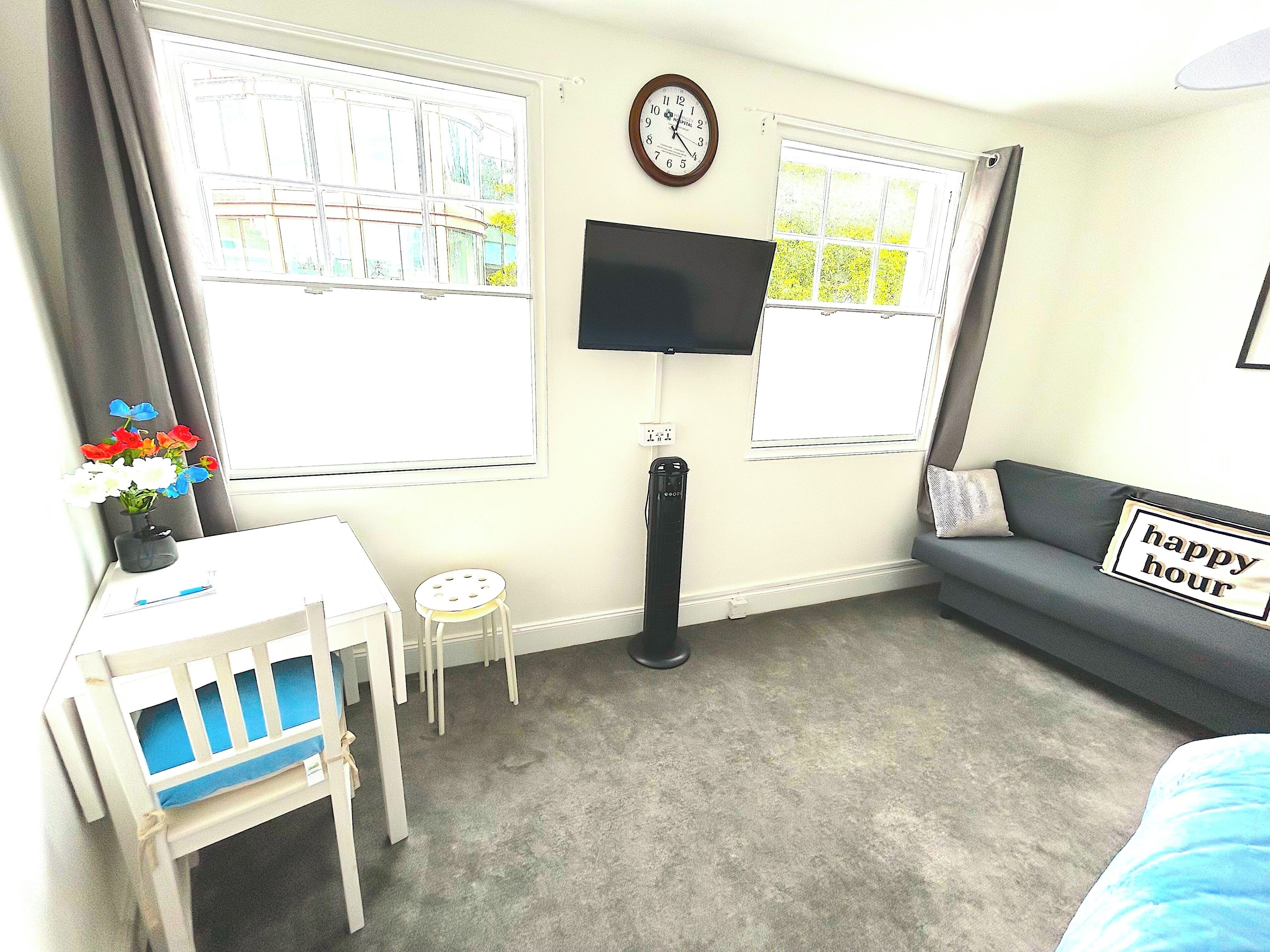 A room with a desk, chair, TV, sofa, and two windows.