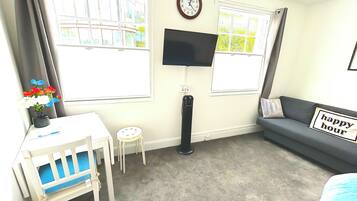 A room with a desk, chair, TV, sofa, and two windows.