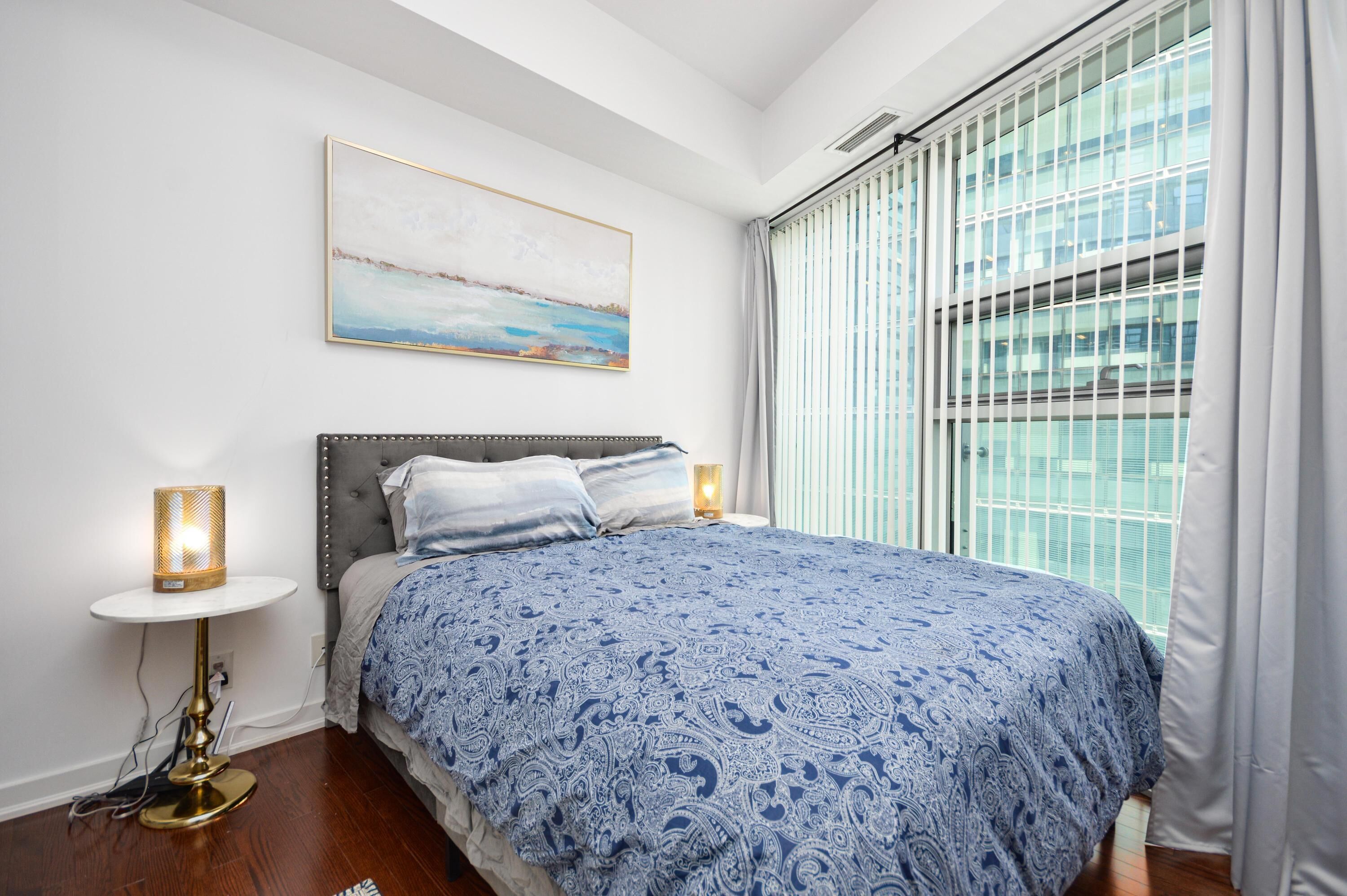 A bedroom with a bed, a bedside table with a lamp, a window with blinds, and a painting on the wall.