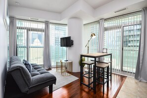A modern living room with a sofa, a dining table with chairs, a TV, and large windows with blinds. - Westmere Suites (Toronto)
