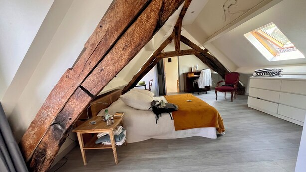 Een slaapkamer met een schuine kap, een bed met witte dekbedovertrek, een houten nachtkastje en een rode stoel.