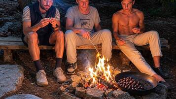 Drie personen zitten bij een kampvuur met een pan eten.