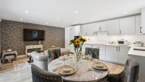 A modern living room with a dining table set for four, a flat-screen TV mounted on the wall, and a kitchen area with white cabinets and appliances. - Calm, Modern Flat with Terrace Near Station - Pass the Keys (Hemel Hempstead)