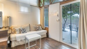 Una habitación con piso de madera, un sofá blanco con cojines estampados, una mesita blanca y un ventanal corredero que da a un espacio exterior.