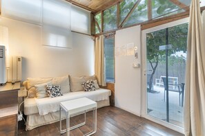 A room with a wooden floor, a white sofa with patterned cushions, a small white table, and a sliding glass door leading to an outdoor area. - Cabañita rústica con piscinas cerca de A Coruña (Peiraio)
