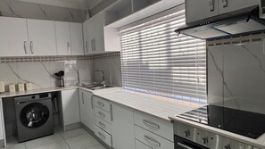 A modern kitchen with a built-in washing machine, stainless steel appliances, and a window with blinds. - SAOB PROPERTY (Germiston)