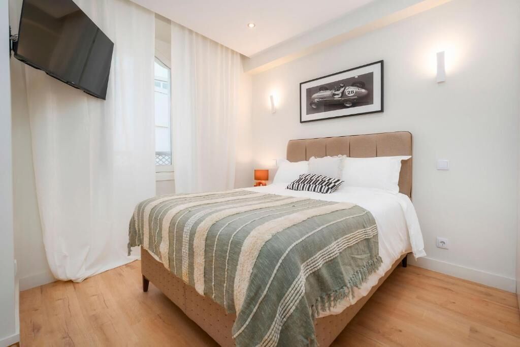 Un dormitorio con cama, televisor montado en la pared, un cuadro enmarcado y una ventana con cortinas.