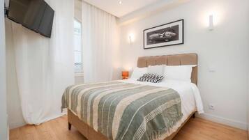 Un dormitorio con cama, televisor montado en la pared, un cuadro enmarcado y una ventana con cortinas.