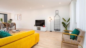 A modern living room with a yellow sofa, a TV on a white stand, a dining area with chairs, and a large plant.