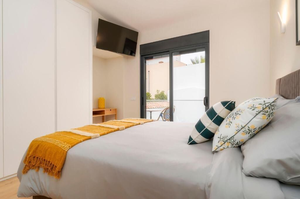 Een slaapkamer met een bed, een aan de muur bevestigde televisie, een balkon met uitzicht, en een kleine tafel met een geel voorwerp.
