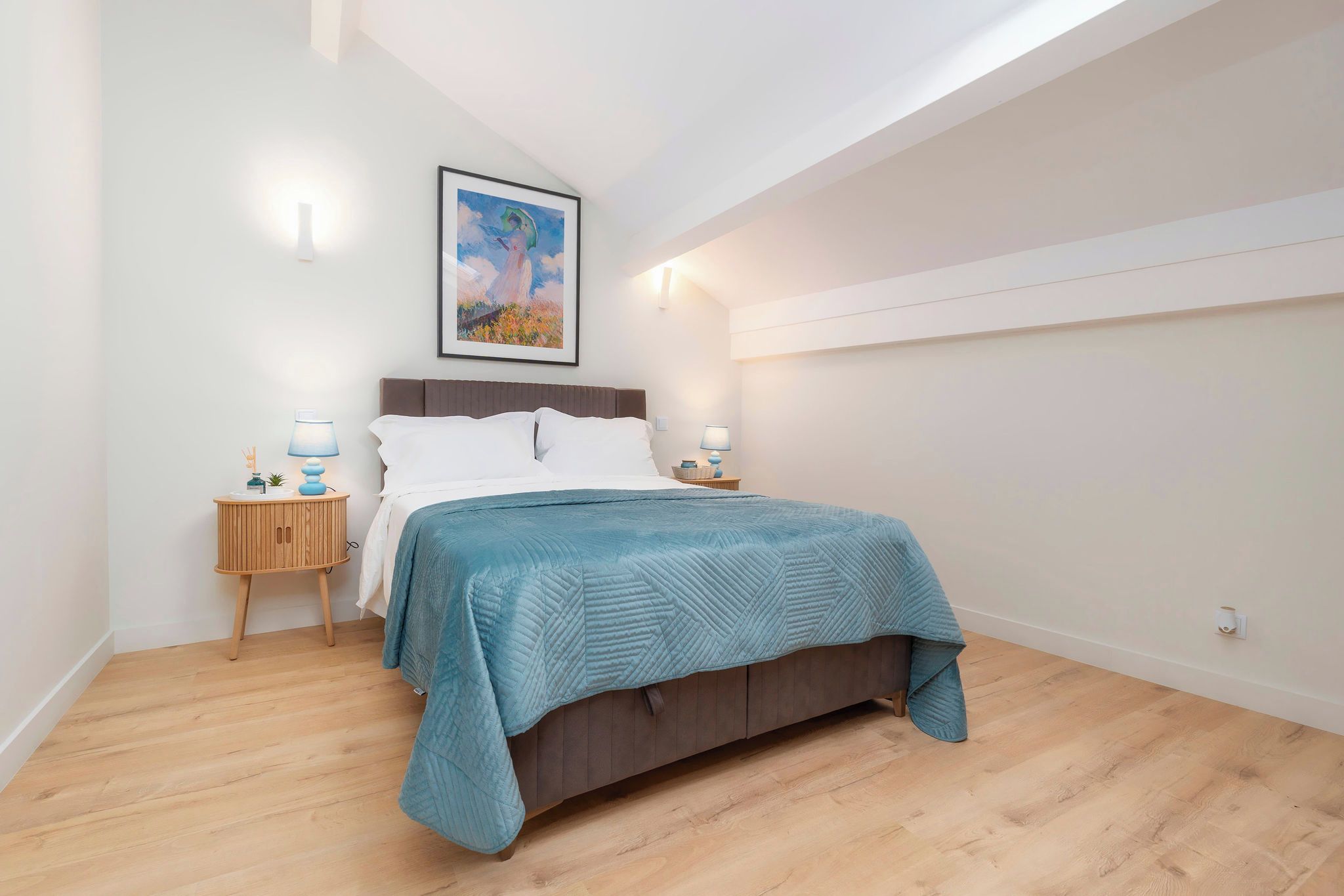 A bedroom with a bed, two bedside tables, a wall-mounted lamp, and a painting.