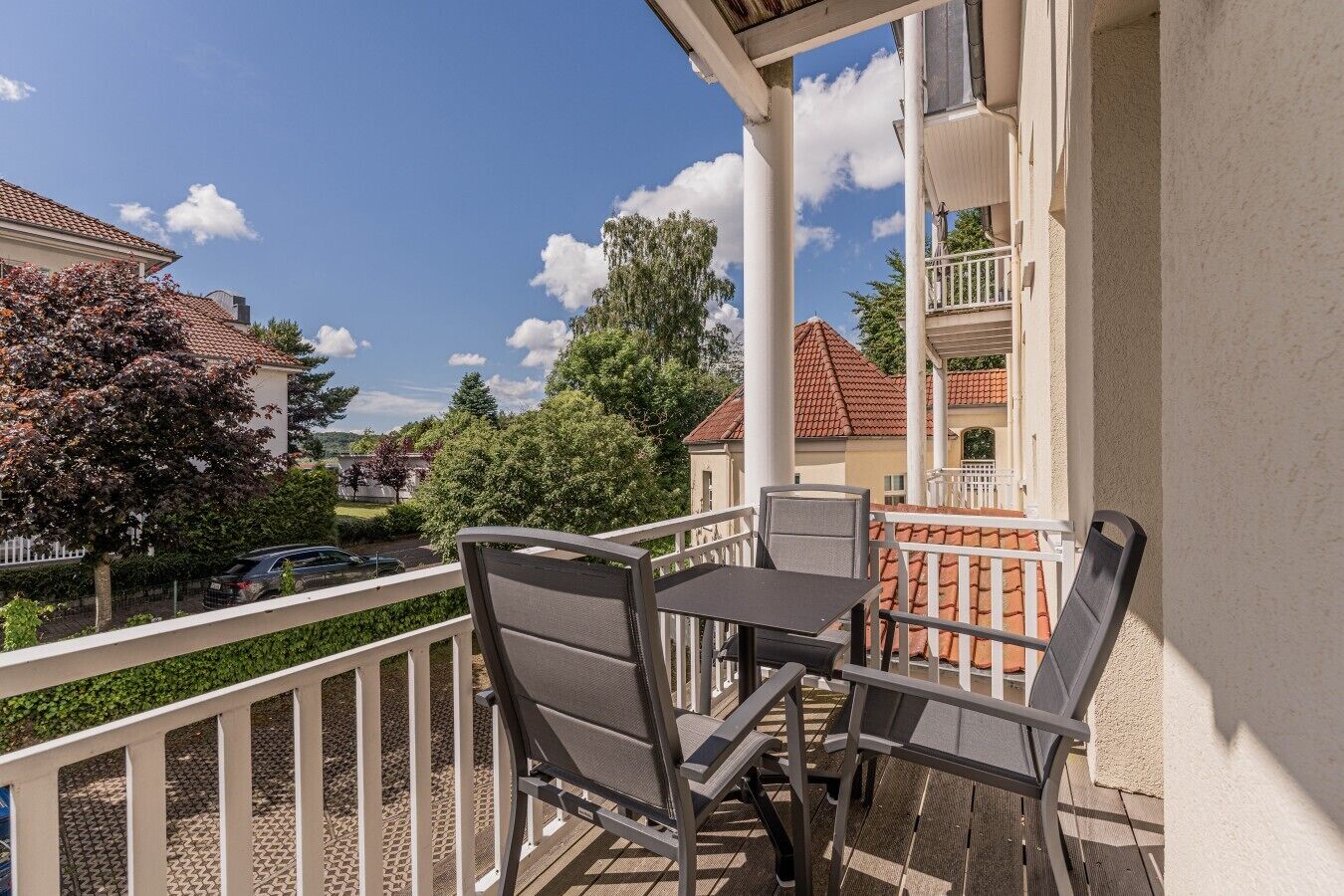 A balcony with a table and chairs, a white railing, and a view of neighboring houses and trees.