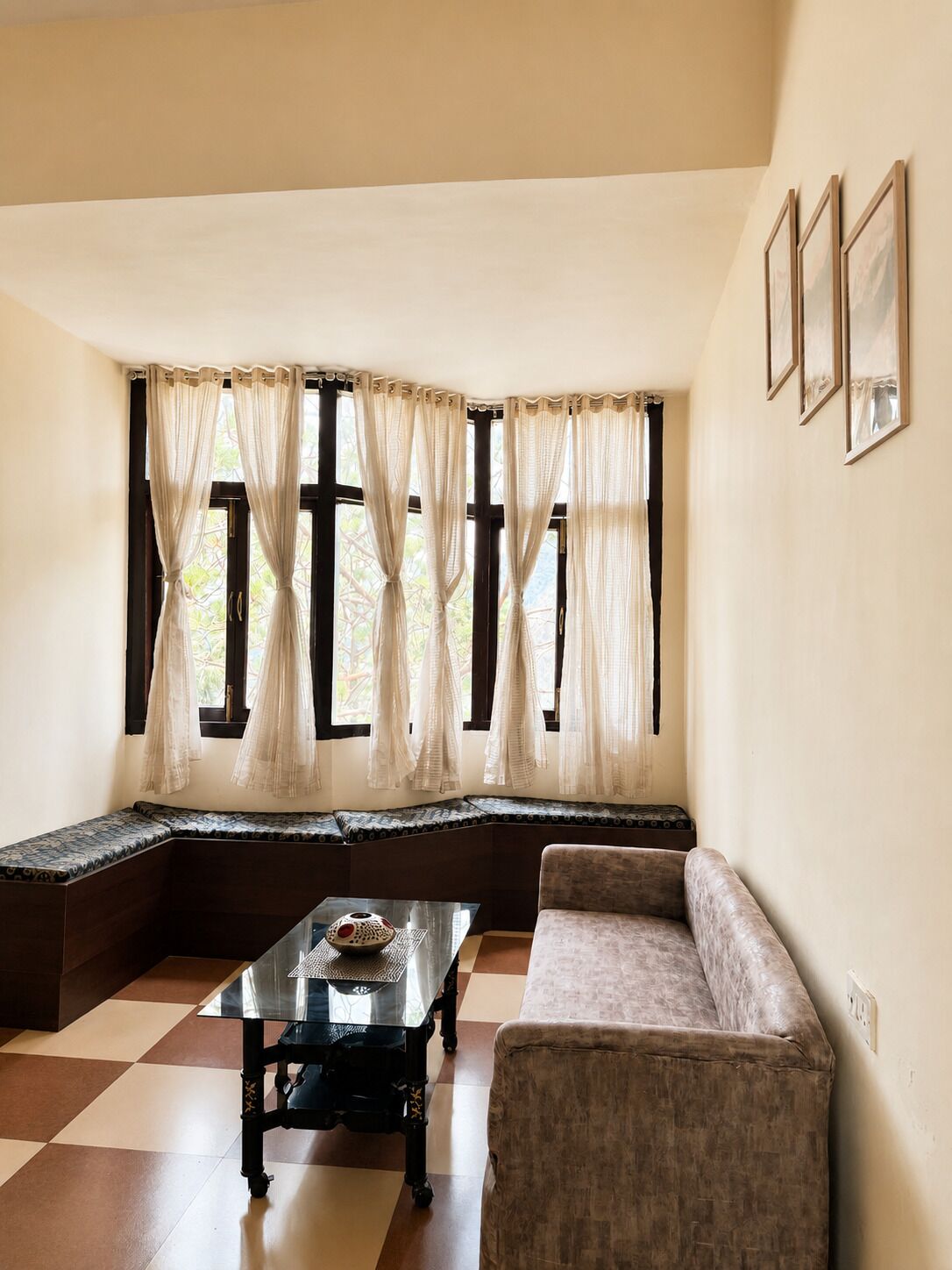 A room with a checkered floor, a sofa, a glass table with a decorative item, a window with curtains, and framed pictures on the wall.