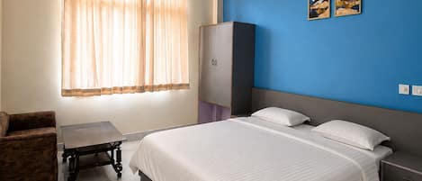 A hotel room with a bed, a desk, a chair, a wardrobe, a ceiling fan, and two paintings on the wall.