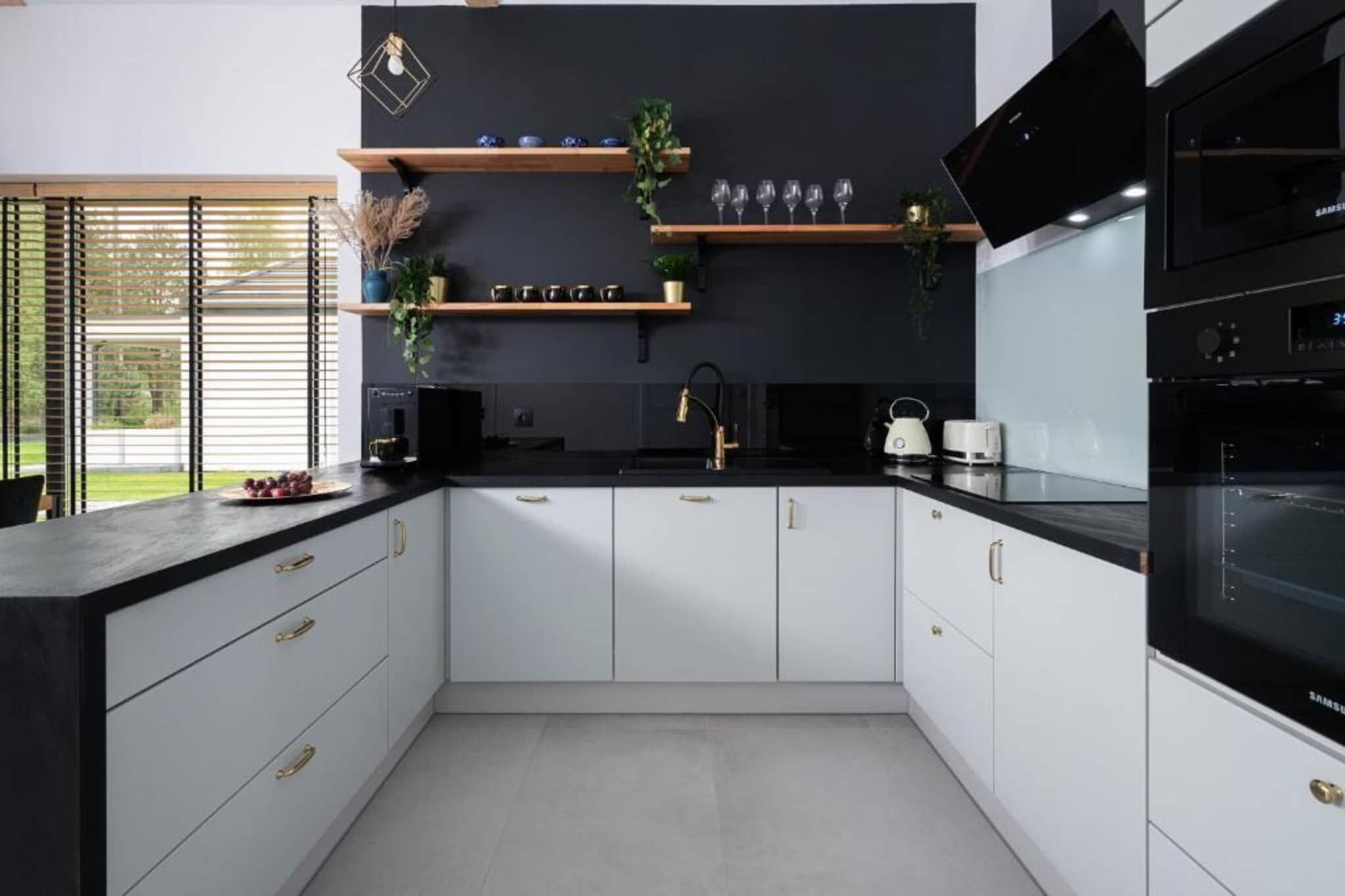 Une cuisine moderne avec des armoires blanches, des comptoirs noirs et des appareils électroménagers en acier inoxydable. On y trouve des étagères en bois ornées d’objets décoratifs et une fenêtre avec des stores.