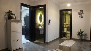 A hallway with a white cabinet, a plant, a mirror with a light, a phone, and a tiled floor. - Cozy/Peaceful/Near Maadi/Ring Road/Gated Compound (Cairo Governorate)