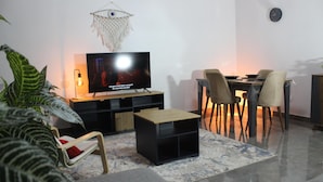 Una sala de estar con televisor de pantalla plana, una mesa de comedor con sillas, una pequeña mesa de centro y un adorno colgante en la pared.