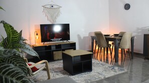 A living room with a flat-screen TV, a dining table with chairs, a small coffee table, and a decorative wall hanging. - Cozy/Peaceful/Near Maadi/Ring Road/Gated Compound (Cairo Governorate)