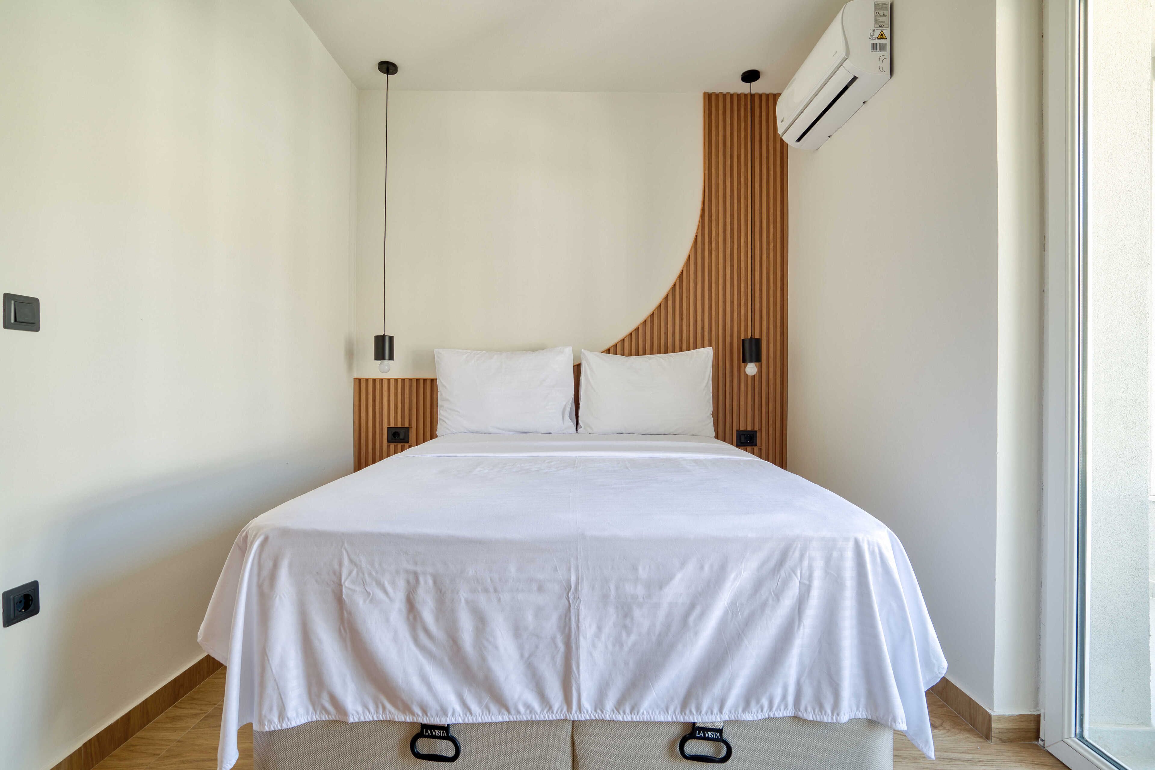 A modern hotel room with a bed, a wooden headboard, a wall-mounted air conditioner, and a window.