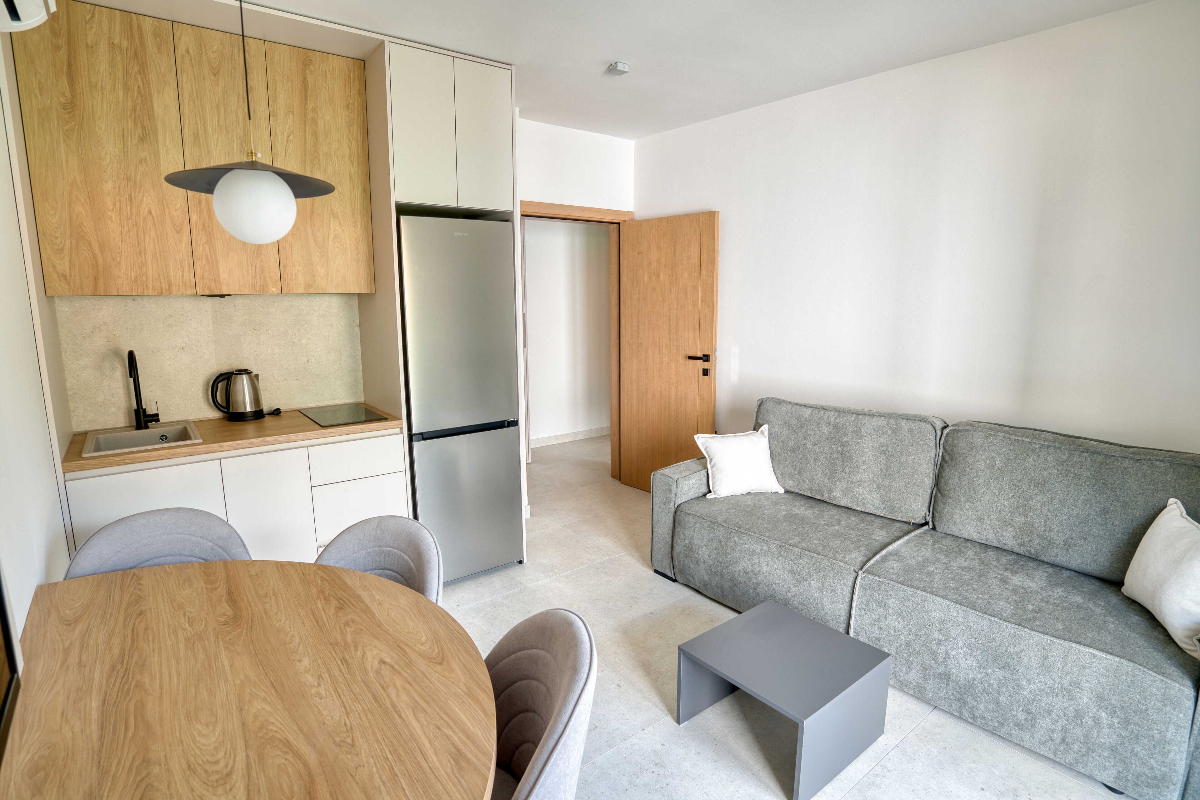 Ein modernes Wohnzimmer mit grauem Sofa, einem Holzesstisch und einer Küchenzeile mit Kühlschrank und Spüle.