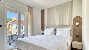 A hotel room with a large bed, a small bedside table, a wall-mounted light, and a sliding glass door leading to an outdoor area. - Mont Residence (Budva)