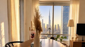 A dining area with a wooden table, a vase with dried plants, a painting, and a view of a cityscape with tall buildings. - Luxury Downtown Skyline Suite Burj Khalifa View (Dubai)