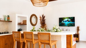A modern kitchen with a central island, wooden chairs, a hanging light fixture, a television, and decorative plants. - Casa Melissa 2BR Pool Villa by TVM (Ubud)