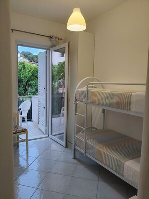 Une pièce avec des lits superposés, une chaise, un luminaire suspendu et une porte-fenêtre coulissante donnant sur un espace extérieur avec une chaise et un arbre.