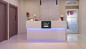 A reception desk with a sign indicating it's company-serviced, a potted plant, a small statue, and a window above the door. - Deluxe Cloud Stay (Guwahati)