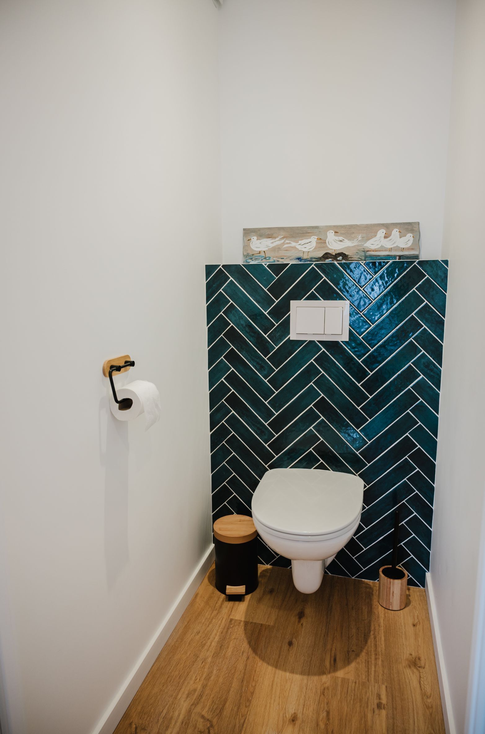 Un baño con inodoro blanco, suelo de madera, pared con diseño de espiga y un pequeño cubo de basura.